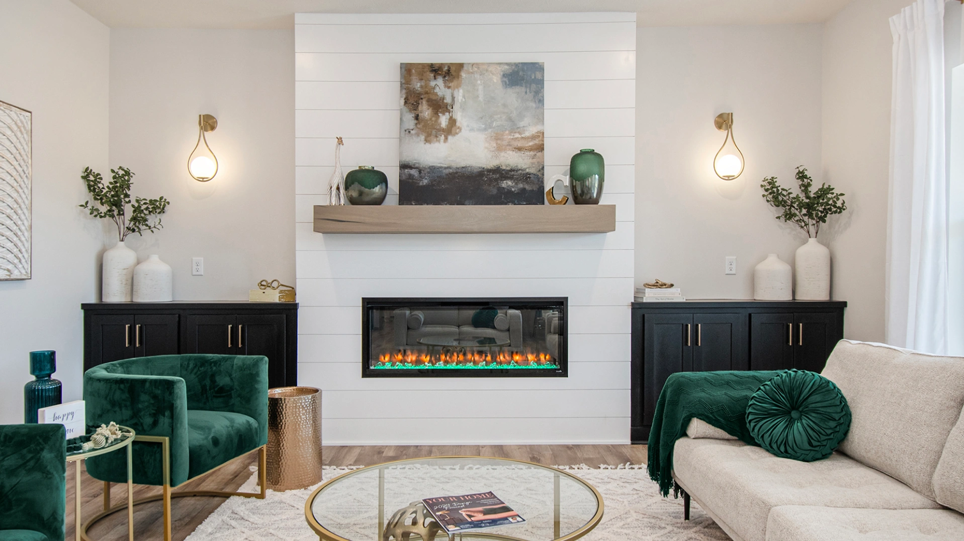 A living room with a fireplace, two green velvet chairs, and a beige sofa