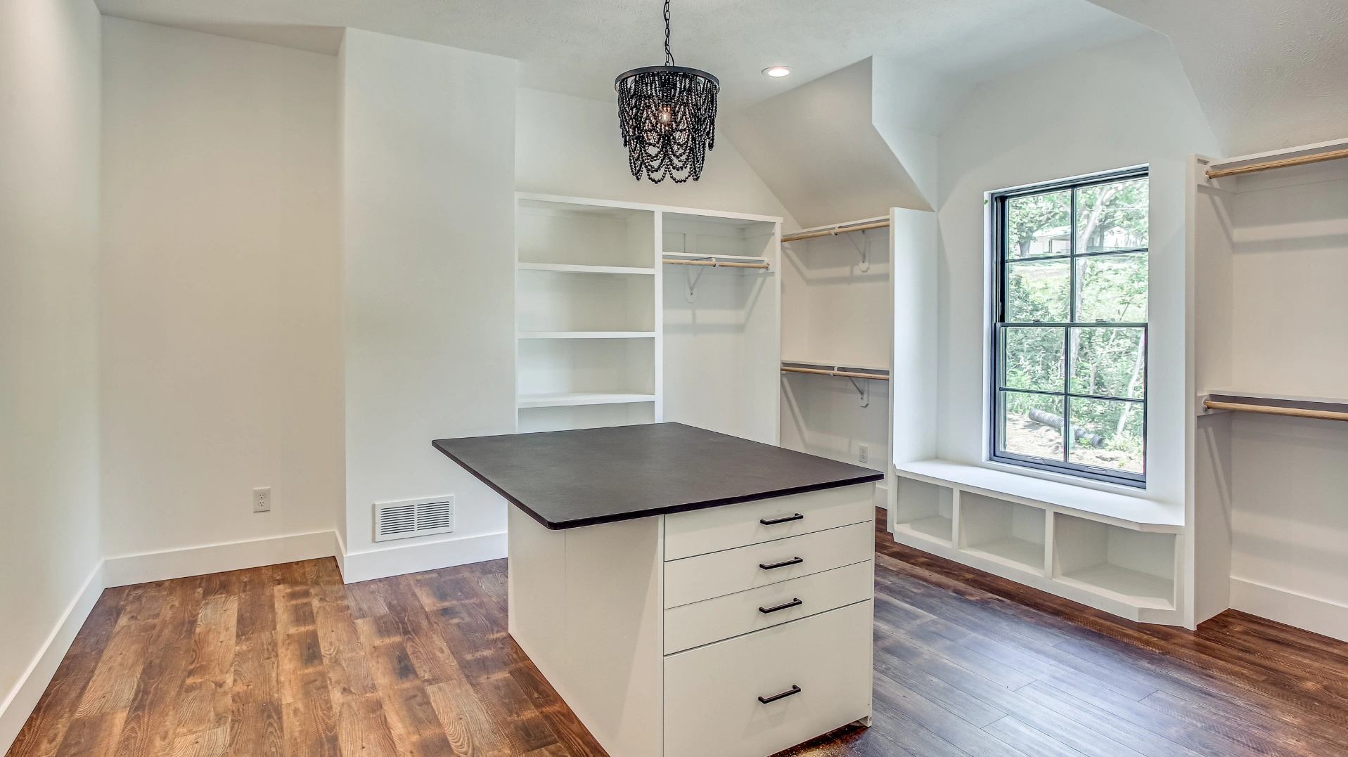 A white closet space with a dark wood floor, a central island with drawers, and shelves surrounding a window